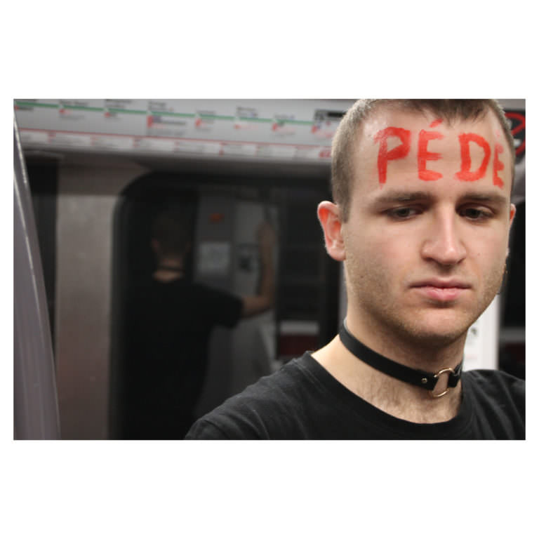 Eine Person, die in der U-Bahn steht und auf der Stirn mit roten Buchstaben das Wort "Pédé" (Schwuchtel) trägt.